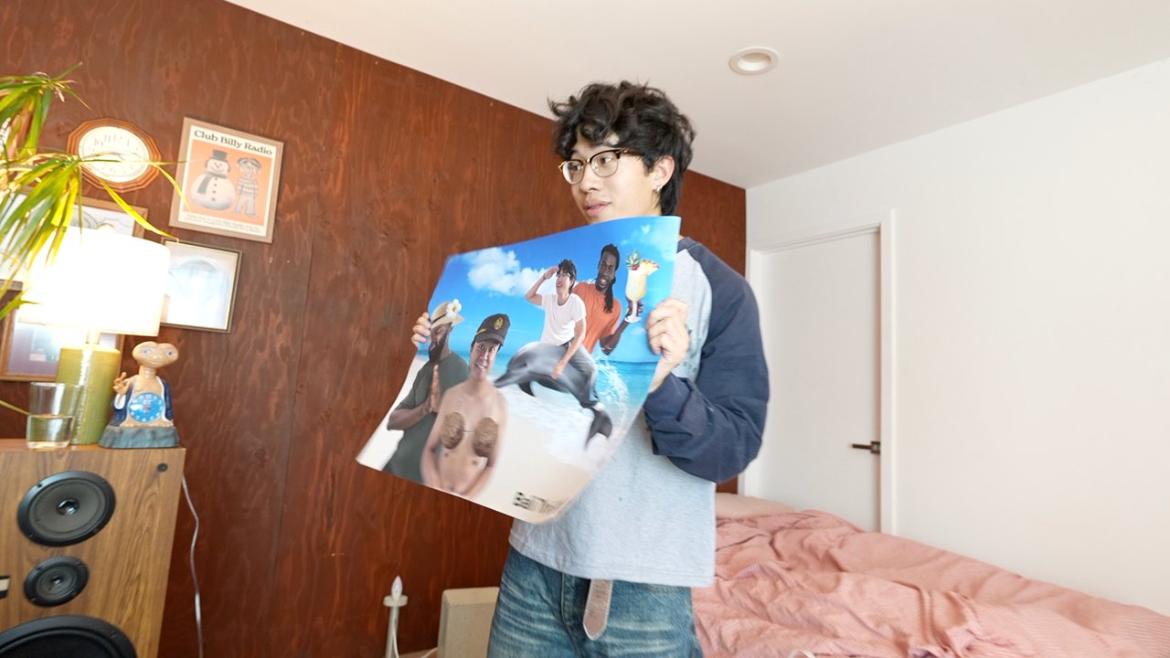 Sneak peek: Billy holding up a printed poster of the crew photoshopped onto a beach with a dolphin